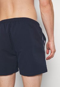 Bañador de color navy con cintura elástica, bolsillo lateral de malla y franjas blancas en los lados. Tejido suave y ligero.
