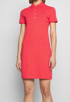 Vrouw draagt een koraalkleurige korte mouw polo jurk met knopen en een klein logo op de borst, staand tegen een effen achtergrond.