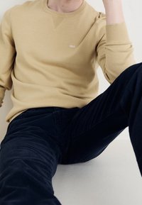 Maglione beige con scollo rotondo testurizzato e piccolo logo sul petto, abbinato a pantaloni in velluto a coste blu scuro. Taglio semplice e rilassato.