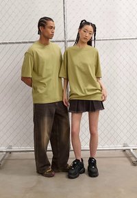 Olive green oversized t-shirts, paired with dark jeans and a black skirt. The setting features a chain-link backdrop with a concrete floor.