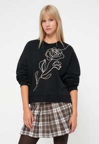 Joven mujer vistiendo un sudadera negra con un dibujo de una rosa beige de líneas y una falda de cuadros marrones pliegue, mirando hacia adelante.
