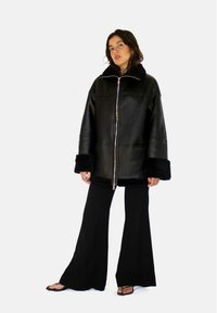Zwarte leren jas met een kraag en manchetten van bont, voorzien van een ritssluiting aan de voorkant, oversized pasvorm en een gladde textuur. Gecombineerd met zwarte broek.