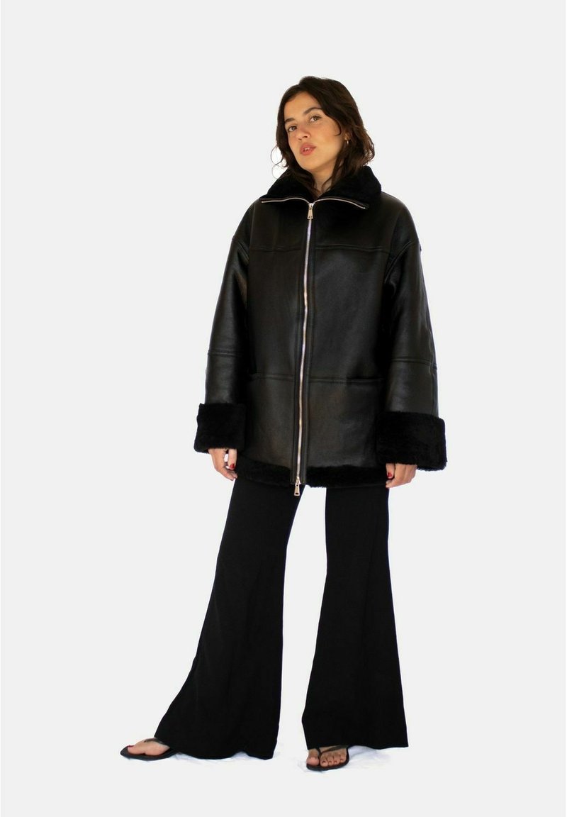 Zwarte leren jas met een kraag en manchetten van bont, voorzien van een ritssluiting aan de voorkant, oversized pasvorm en een gladde textuur. Gecombineerd met zwarte broek.