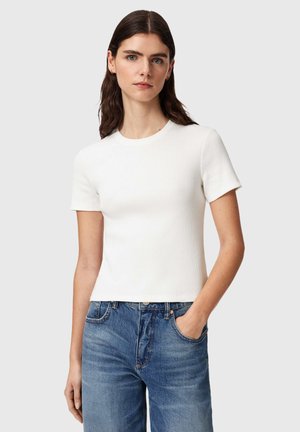 Kobieta o długich ciemnych włosach, ubrana w biały, prążkowany top z krótkim rękawem i niebieskie dżinsy, stoi z jedną ręką w kieszeni.