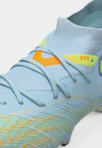 Ανοιχτό μπλε ποδοσφαιρικό παπούτσι με ψηλό κολάρο στον αστράγαλο, κίτρινες και πορτοκαλί λεπτομέρειες και ανοιχτό μπλε κορδόνια σε υφή επιφάνειας.