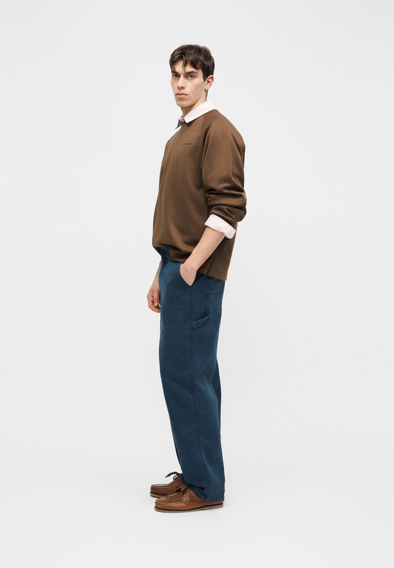 Jeune homme debout de profil portant un sweat-shirt marron sur une chemise blanche, un pantalon bleu ample et des chaussures en cuir marron sur fond uni.