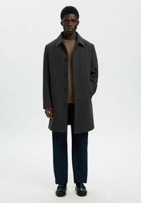 Pelēkā rūtainā jakā ar norādītu apkakli, pogu aizdari un sānu kabatām, uzvilkta pāri gaiši brūnai sporta jaku un tumši zilām biksēm.