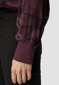 Bordeaux langemouwen shirt met een gestructureerd, abstract zwart patroon op de mouw. Omgeslagen zoom in effen bordeaux. Zwarte broek zichtbaar aan de onderkant.