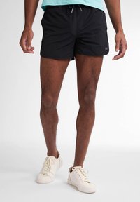 Pantaloni scurți negri de baie, confecționați dintr-un material ușor, cu un brâu elastic și șnur. Asortați cu adidași albi.