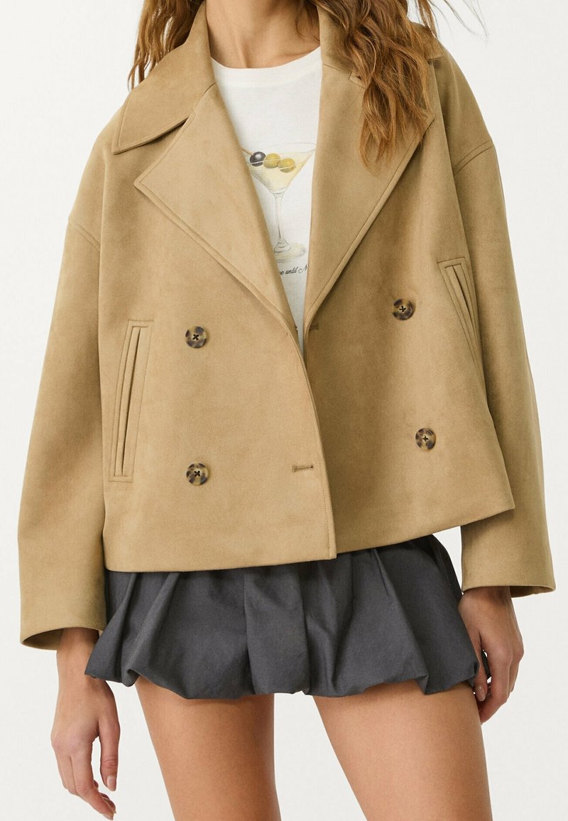 Veste croisée couleur beige en tissu doux avec de grands revers, deux poches latérales et des boutons ronds à motif. Portée avec un t-shirt graphique et une jupe.