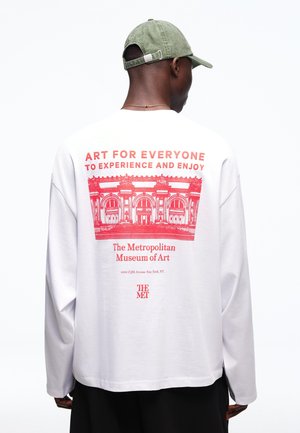 Man draagt een wit shirt met lange mouwen met een rood afdruk van de gevel van het Metropolitan Museum of Art en tekst die kunsttoegankelijkheid op de achterkant promoot.