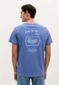 Tricou din bumbac albastru cu un design pe spate, având text grafic alb "MÂNCAREA ZILEI" și o ilustrație cu pește, etichetat "Pește proaspăt din CANADA ESTICĂ."