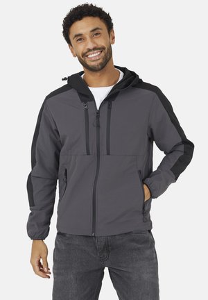 Mężczyzna uśmiechający się, ubrany w szarą i czarną bluzę z kapturem na zamek błyskawiczny, z kieszeniami na zamek i elastycznymi mankietami, w połączeniu z ciemnoszarymi dżinsami.