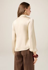 Cremfarbene Bluse mit langen Ärmeln und hohem Kragen; glatte Textur und leicht gepuffte Ärmel. Kombiniert mit braunen Hosen und einem roten Armreif.