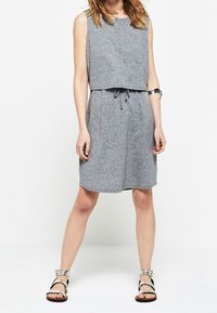 Robe sans manches grise avec une coupe ample, taille à cordon et ourlet au genou. Portée avec des sandales noires et argentées. Tissu texturé.