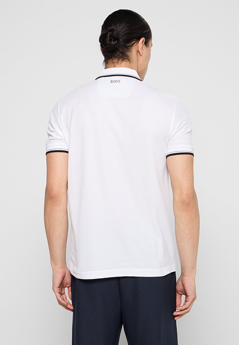 Camiseta polo blanca con ribete negro en el cuello, mangas cortas y un detalle de logo en la parte posterior del cuello. Textura suave, ajuste estándar, diseño casual.