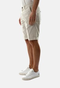 Pantaloncini di cotone beige con taglio dritto, tasche laterali e orlo piegato, abbinati a sneakers bianche, mostrati di lato.