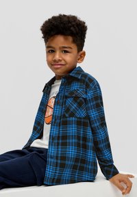 Karierte Flanellhemd in Blau und Schwarz mit Knopf-Taschen, über einem weißen Grafik-T-Shirt getragen, kombiniert mit marineblauen Hosen.