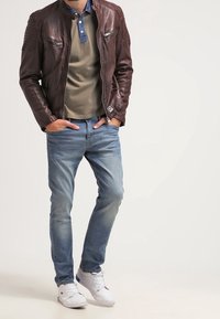 Veste en cuir brun avec poches zippées, chemise vert olive avec col bleu, jeans bleu clair et baskets blanches. Se tenant contre un fond neutre.