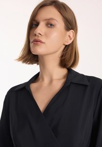Czarna koszula ze zwężającym się fasonem, z kołnierzykiem, o gładkiej fakturze i delikatnym połysku. Materiał układa się miękko wokół obojczyka.