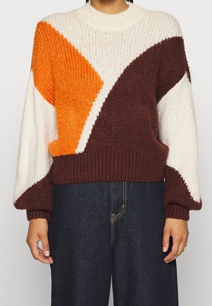 Personne portant un pull en grosse maille avec des blocs de couleurs géométriques blanc, orange et marron, associé à un pantalon taille haute bleu foncé.