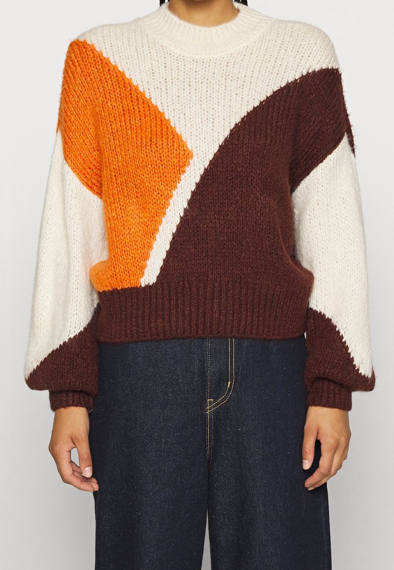 Personne portant un pull en grosse maille avec des blocs de couleurs géométriques blanc, orange et marron, associé à un pantalon taille haute bleu foncé.