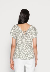 Vrouw met lang, donker haar, gekleed in een grijs-witte bloemenblouse met korte mouwen en een geknoopte V-hals aan de achterkant, in combinatie met witte broek, met haar rug naar de camera gericht.