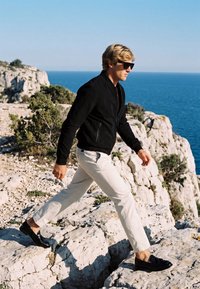 Homme en veste noire, pantalon blanc et lunettes de soleil marchant sur des falaises rocheuses près de l'océan sous un ciel bleu clair.