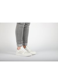 Weiße Sneaker mit glatter Oberfläche und hellgrauen Wildlederakzenten; runde Zehenpartie, flache Sohle und Schnürdesign. Getragen mit hochgekrempelten grauen Jeans.