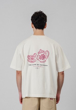 Mand iført offwhite T-shirt med lyserød tekande og tekop grafik og teksten "The House of Pockies" på ryggen, beige bukser, vendt væk.