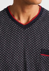 Camiseta de cuello en V color navy que presenta un patrón de diamantes en blanco y rojo, con un bolsillo y acentos rojos contrastantes en el cuello y el bolsillo.
