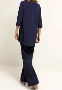 Mörkblå tunika med trekvartslånga ärmar, som har en rak skärning och sidoflikar, kombinerad med matchande vida byxor. Slät tygtextur.