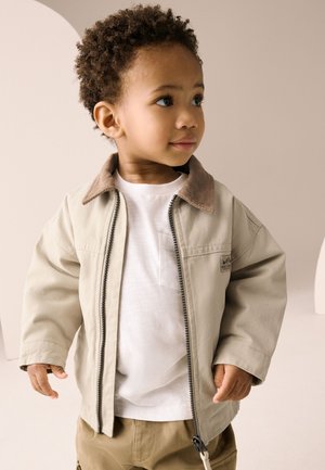Beige jakke med lynlås og brun fløjlsflip, kombineret med en hvid skjorte. Har en afslappet pasform og synlige sømdetaljer.
