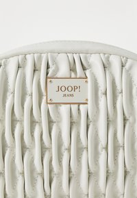 Fodrad vit läderhandväska med en guldfärgad märkesetikett som läser "JOOP! JEANS." Har texturerade, vågiga ytor och dekorerad sömnad.