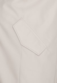 Vaaleanbeige kangas, jossa on kolmion muotoinen päällyspocketsi, jossa on näkyvät ompelu-yksityiskohdat. Sileä pinta ja minimalistinen muotoilu.