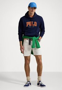 Μπλε σκούφο φούτερ με το λογότυπο «Polo» σε πορτοκαλί, λευκά σορτς, πράσινο πουλόβερ δεμένο στη μέση, μπλε καπέλο και μπλε αθλητικά παπούτσια με λευκές σόλες.