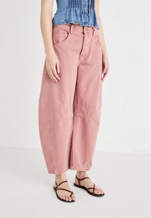 Mujer con vaqueros de pierna ancha en color rosa polvoriento, camiseta sin mangas azul y sandalias planas marrones con tiras, de pie frente a un fondo blanco liso.