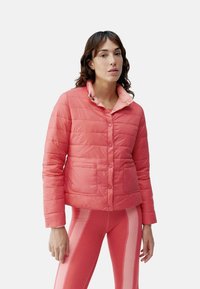 Pūšamā īsā jaka korallā, ar pogu priekšpusi, divām priekšējām kabatām un kontrastējošu apkakli. Gluda tekstūra ar izšūtu rakstu.