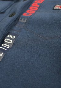 Chemise bleu marine en tissu texturé, avec texte imprimé en rouge et blanc et petit logo de l'union jack sur la zone de la poche poitrine.