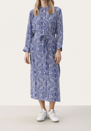 Blau gemustertes Kleid mit Kragen, Knopfleiste, langen Ärmeln und Taillengürtel. Das Outfit wird durch weiße Sneakers abgerundet.