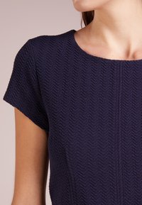 Marinblå klänning med en strukturerad zick-zackmönster, rund halsringning och korta ärmar. Tyget ser mjukt ut med en lätt stretch.