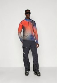 Haut à manches longues avec col zippé, dégradé du rouge au bleu, associé à un pantalon foncé avec poches zippées et logo ; porté avec des chaussures de ski et des lunettes de soleil.