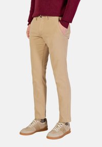 Chinos beiges à coupe ajustée, dotés de poches latérales et d'une fermeture par bouton. Portés avec des baskets gris clair et un pull bordeaux.