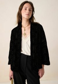 Zwarte fluwelen blazer met geometrisch patroon, losse pasvorm, driekwart mouwen, gedragen over een crèmekleurige kanten top en zwarte broek.