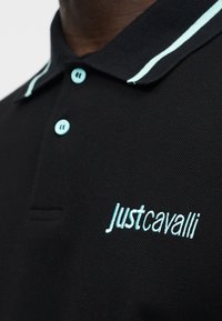 Μαύρο πόλο πουκάμισο με ανοιχτό μπλε κουμπιά, ρίγες στο γιακά και το κέντημα «Just Cavalli» σε ανοιχτό μπλε στο στήθος.