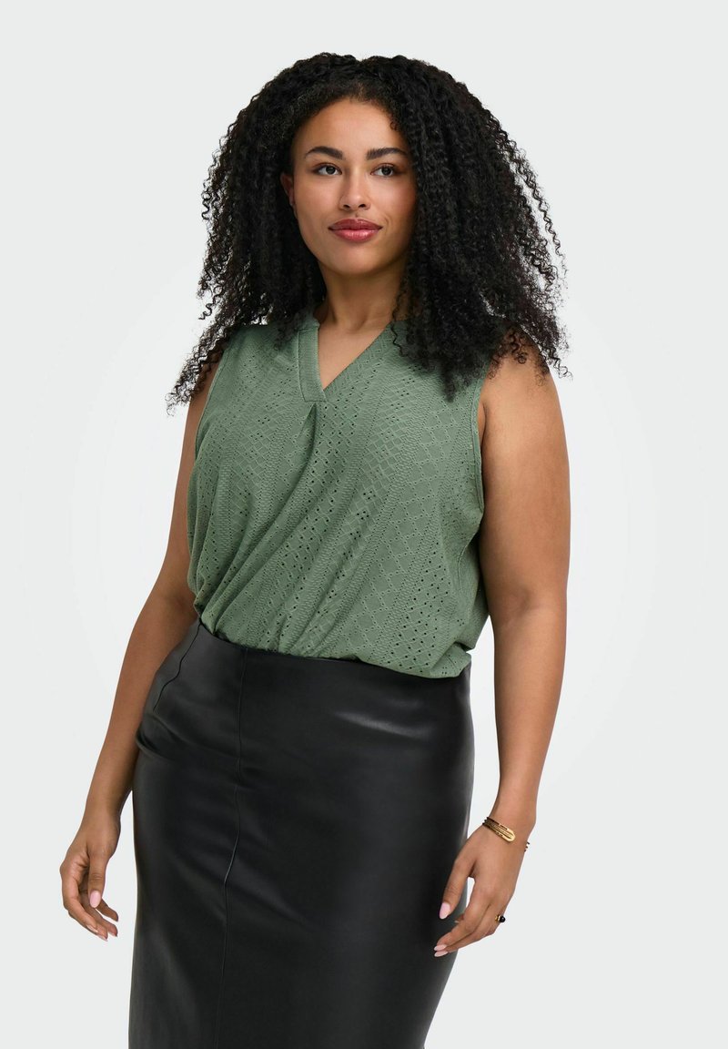 Femme aux cheveux bouclés portant un haut vert sans manches texturé et une jupe en cuir noire, debout devant un fond uni.