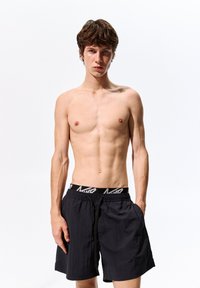 Pantaloni da bagno neri con vita elastica e logo a contrasto. Il tessuto è leggero con una finitura testurizzata.