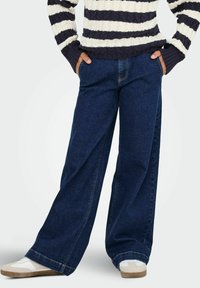 Persona que lleva vaqueros de pierna ancha azul oscuro, zapatillas blancas y un suéter a rayas blanco y negro con las manos en los bolsillos.