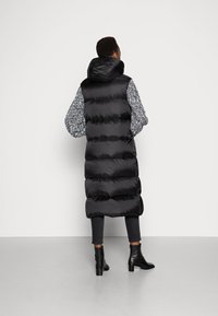 Gilet lungo nero imbottito con cappuccio, caratterizzato da maniche a maglia grigia a motivo, texture trapuntata e lunghezza fino al ginocchio. Stivaletti neri alla caviglia.