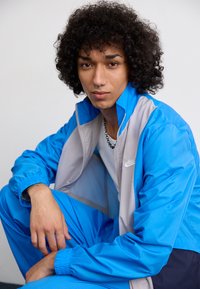Veste de sport bleu et gris en tissu brillant, dotée d'une taille et de poignets élastiques, avec un petit logo sur la poitrine.